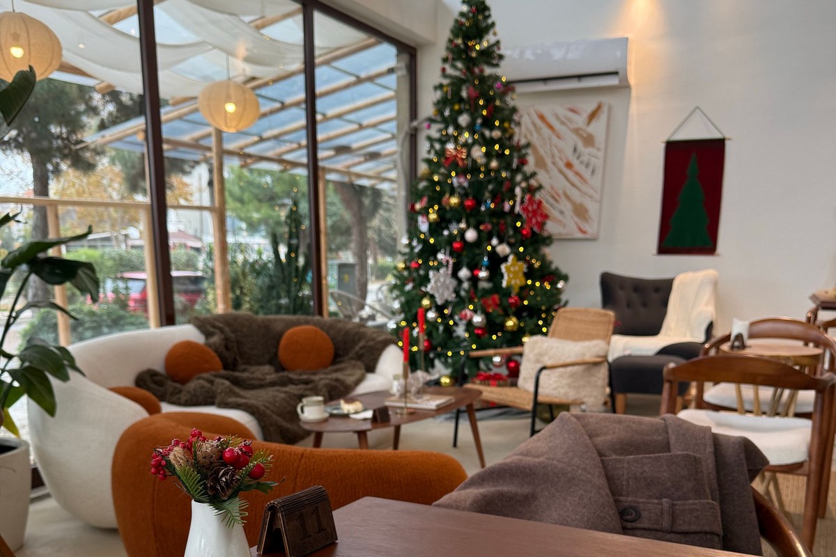 Cozy cafe with a decorated Christmas tree, soft armchairs, and a small holiday flower arrangement on a wooden table.