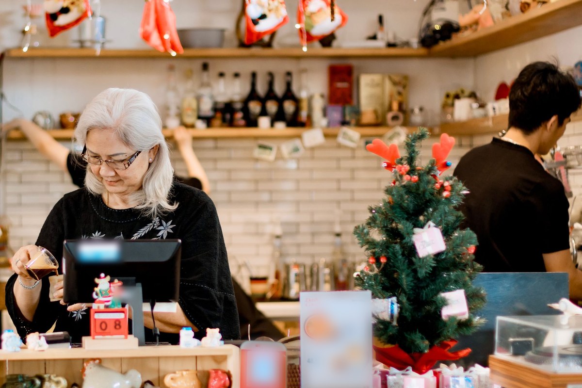 Shop owner working at a holiday-decorated counter beside a small Christmas tree while a coworker prepares orders nearby.