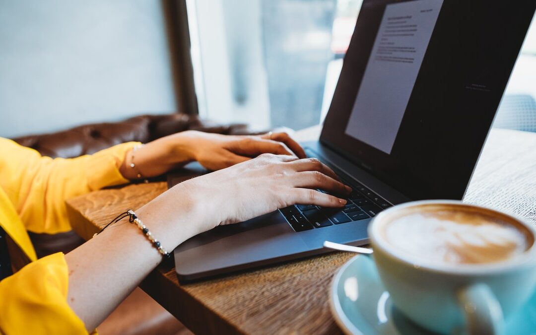 Close-up of a person typing on a laptop at a wooden cafe table beside a cup of coffee